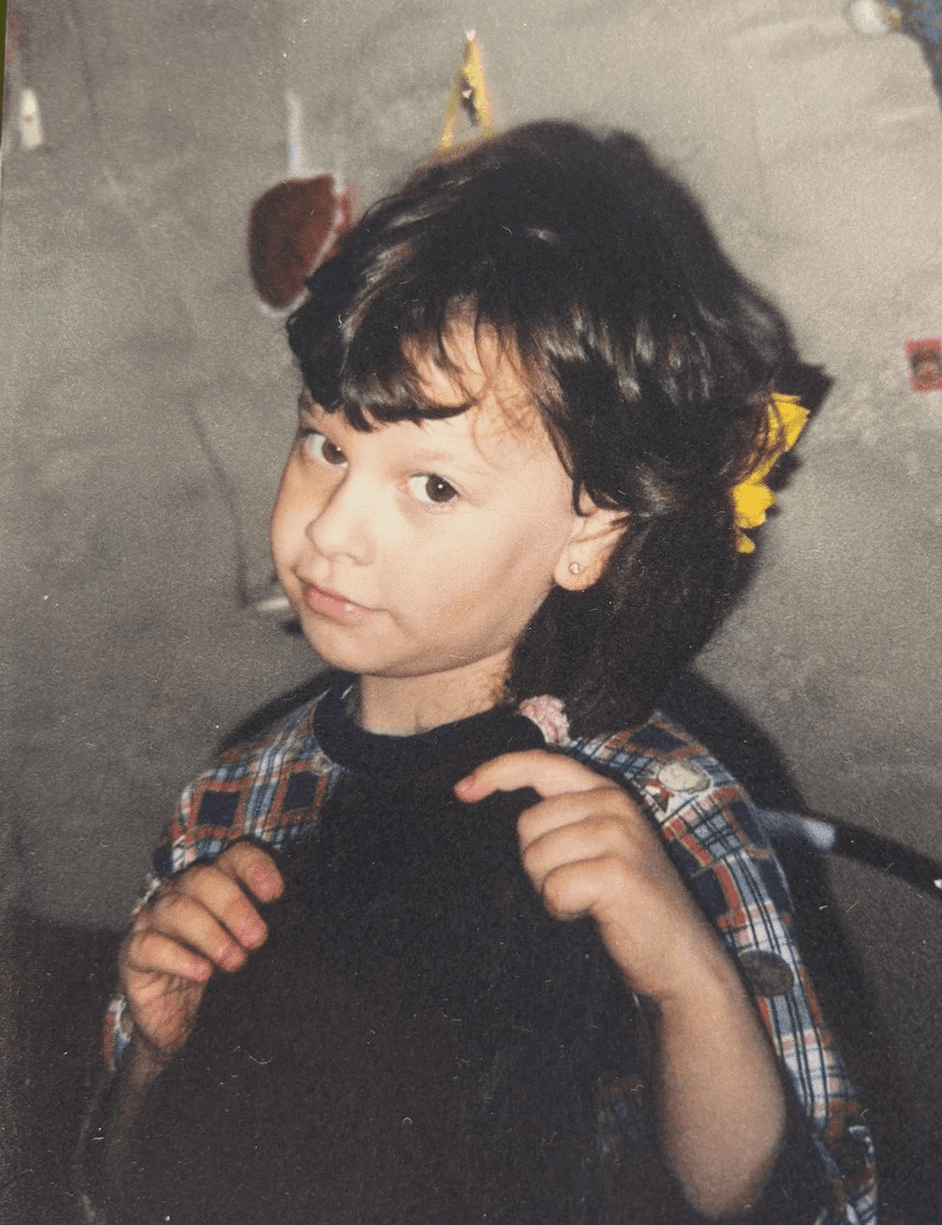 Portrait of a woman with a messy bun and hoop earrings wearing a white paint-splattered T-shirt against a gray background.