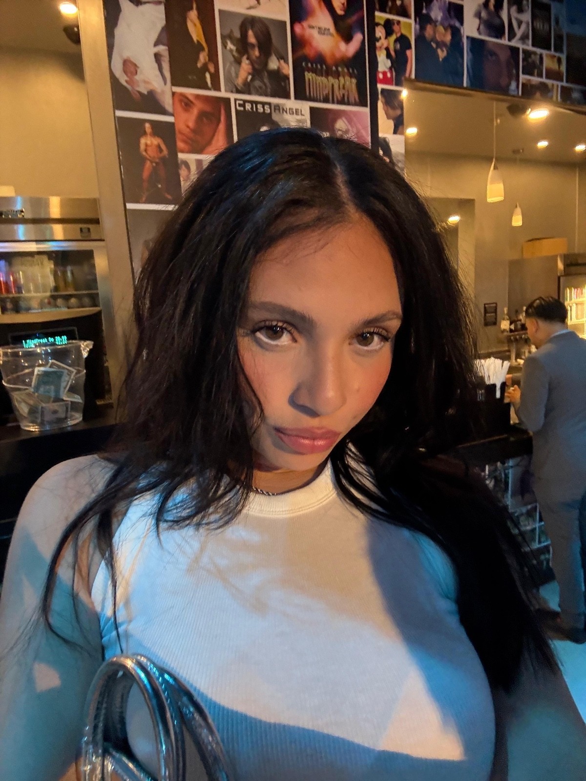 A person with long dark hair and wearing a white top holds a shiny silver purse, standing in a modern, dimly lit room decorated with various photographs on the walls.