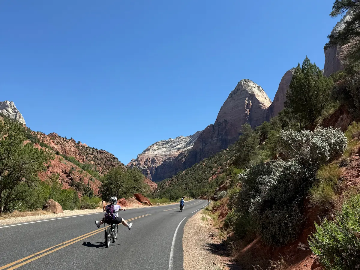 Sara riding an electric bike downhill toward The Narrows in Zion National Park, with Carrie behind her coasting with her feet off the pedals.