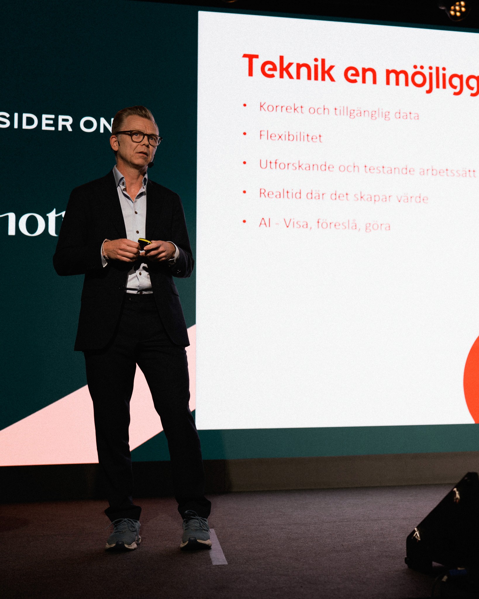 Talare i kavaj på scenen bredvid en presentationsbild med punktlista