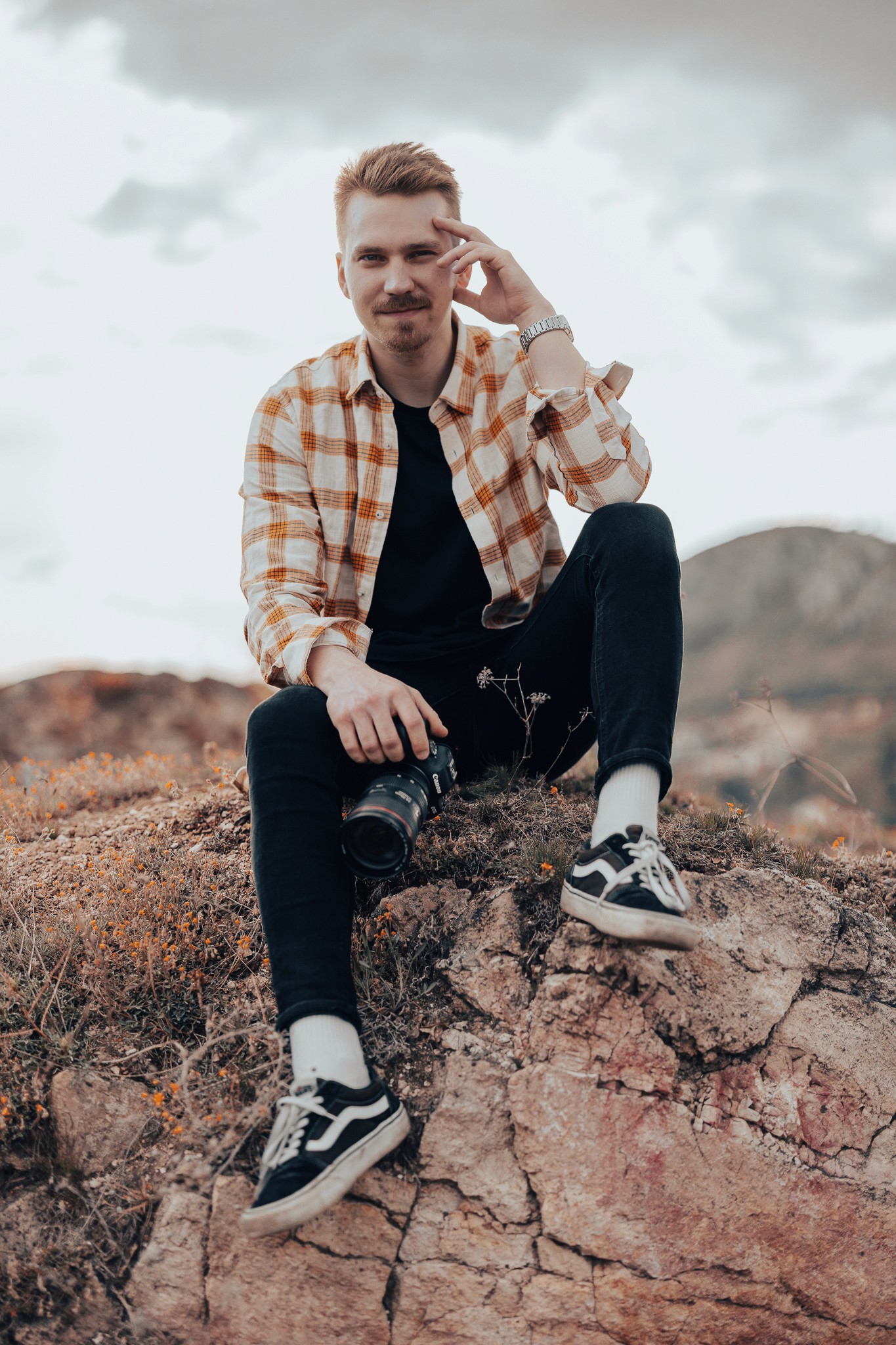Egy fiatal férfi ül egy sziklás dombon, kezében fényképezőgéppel, kockás ingben, fekete nadrágban és sportcipőben.