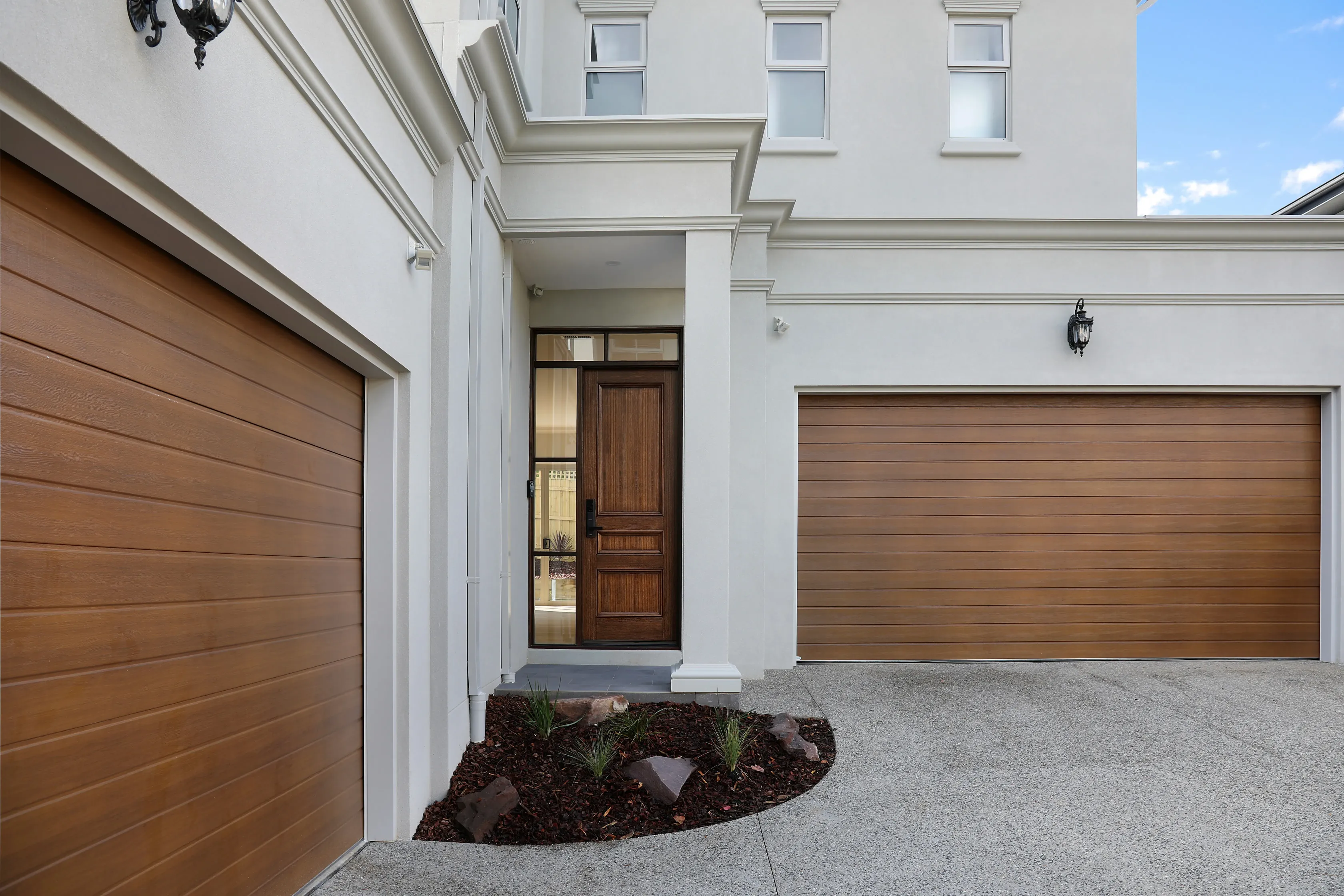 Entry and double timber garage doors of the Glen Waverley Residence, with a render facade, columned portico, panelled timber front door and a mulched garden bed beside the aggregate driveway