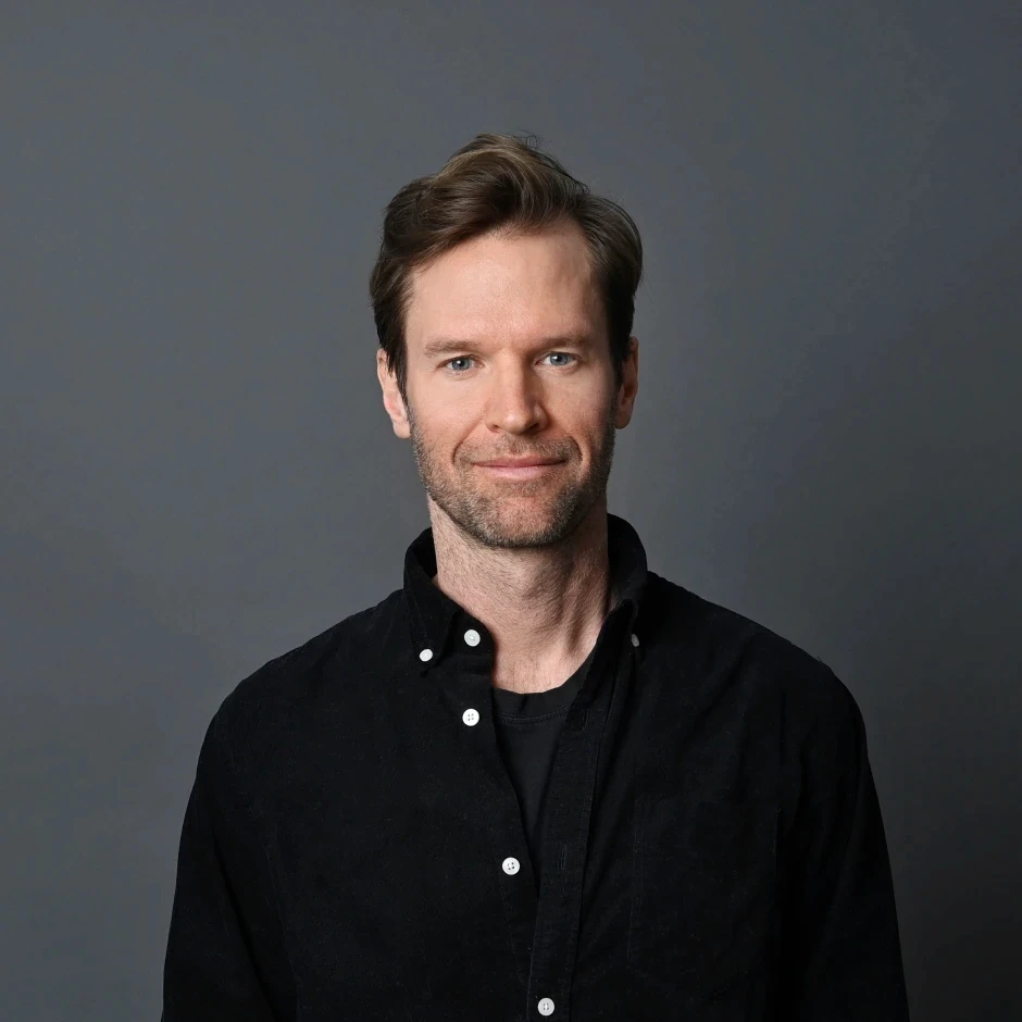 Smiling man with light brown hair wearing a black button-down shirt, looking at the camera.