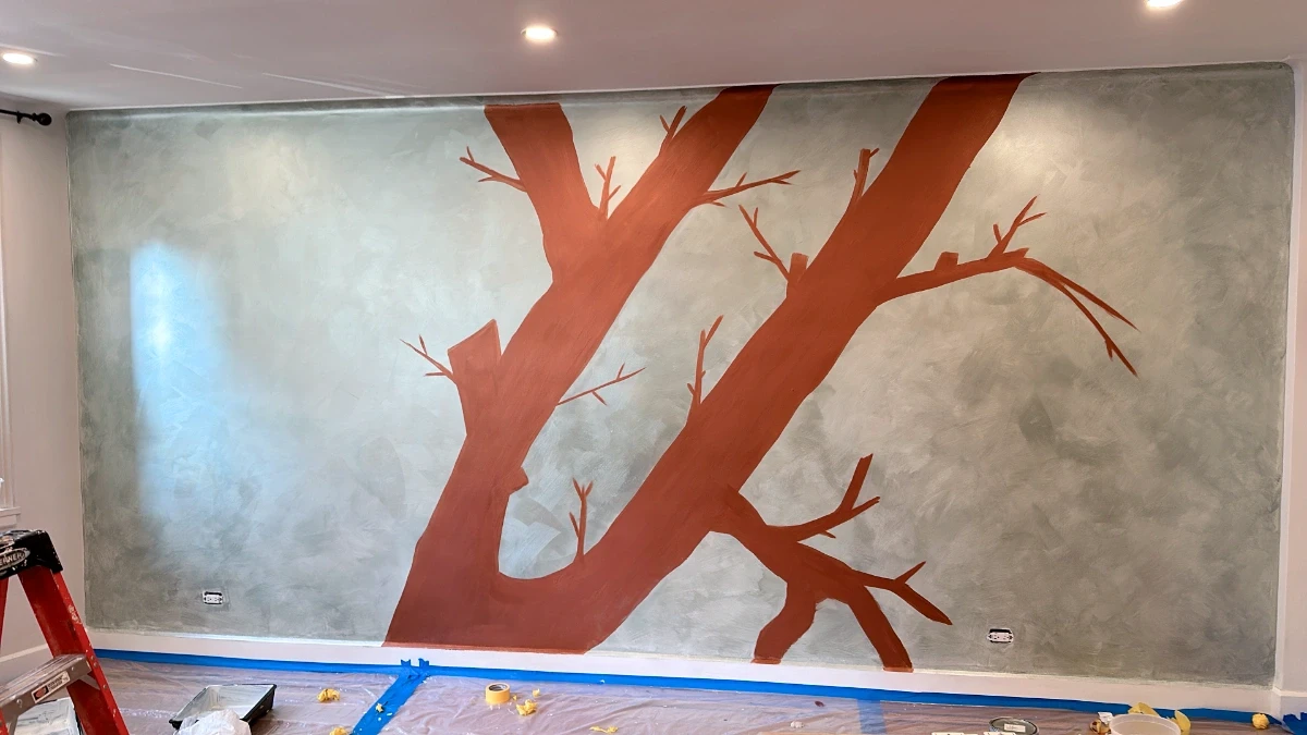 Tree mural in progress on a Venetian plaster wall, featuring hand-painted reddish-brown branches against a soft gray textured backdrop.