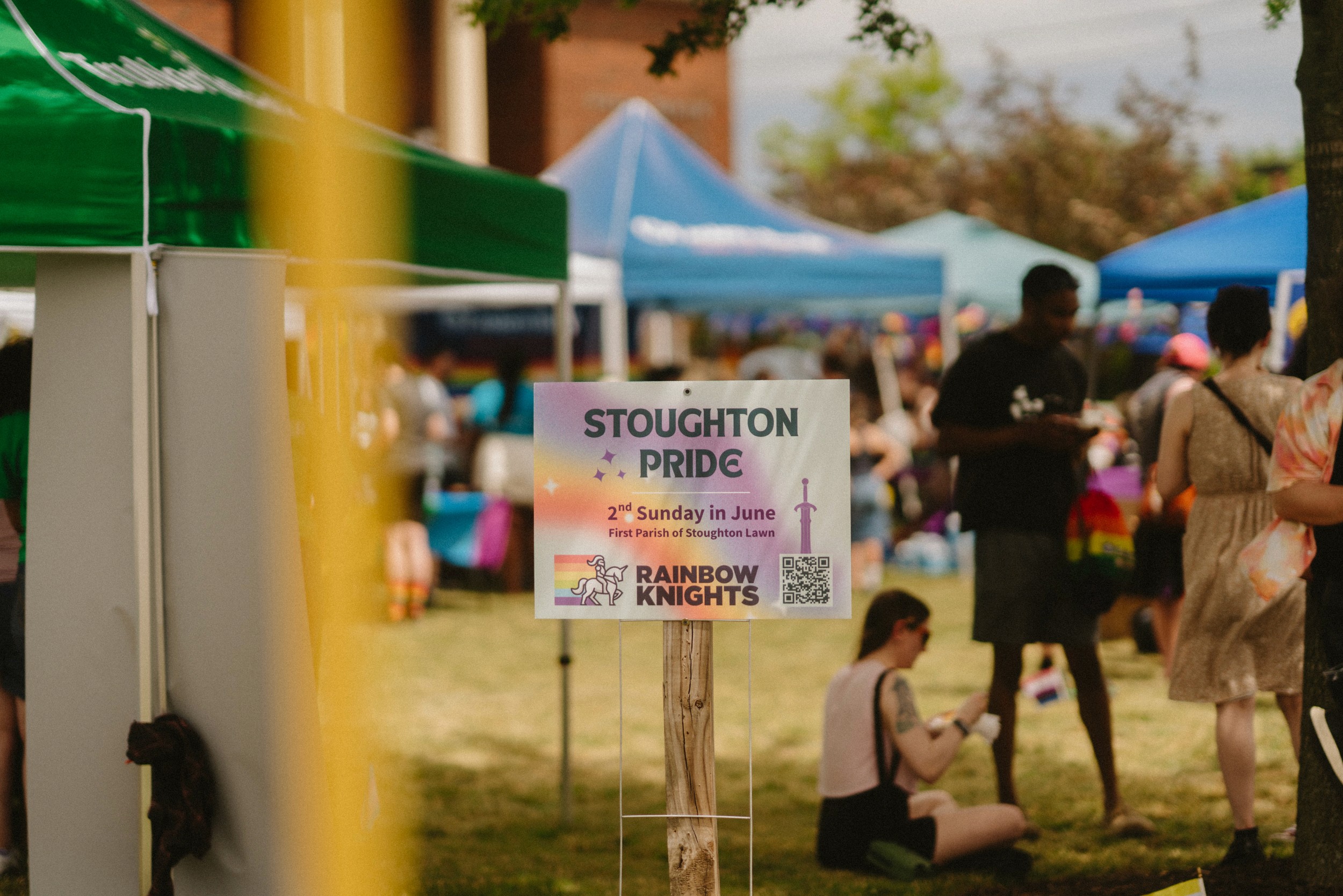 A wooden Stoughton Pride sign with the Rainbow Knights logo stands in front of vendor tents and attendees in motion.