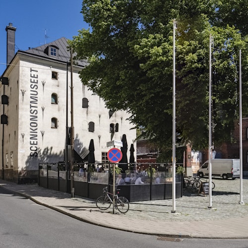 Utomhusområde med sittplatser nära en vit byggnad märkt "Scenkonstmuseet", en "parkering förbjuden"-skylt och cyklar i närheten.