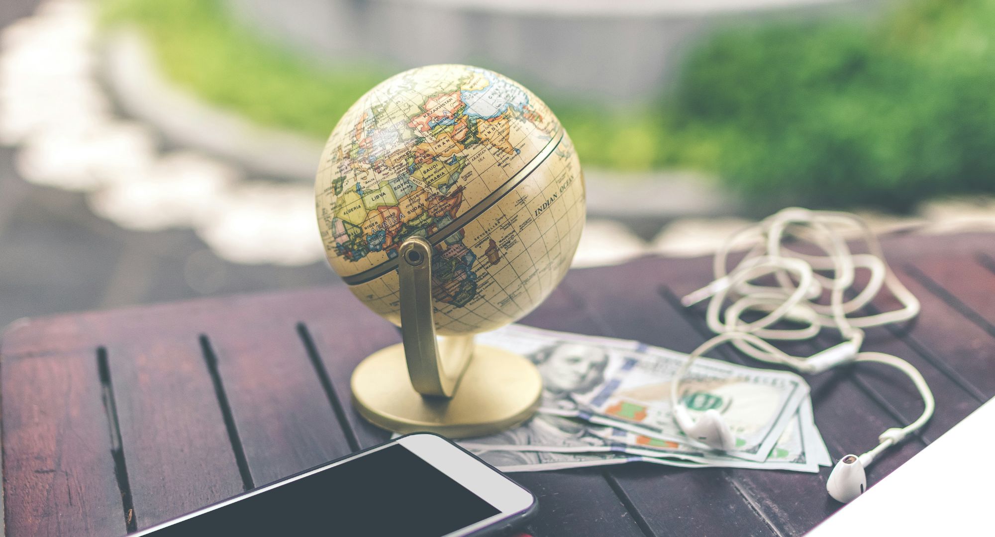 um pequeno globo giratório em uma superfície de madeira junto com um smartphone, um fone de ouvido e duas notas de dólares americanos