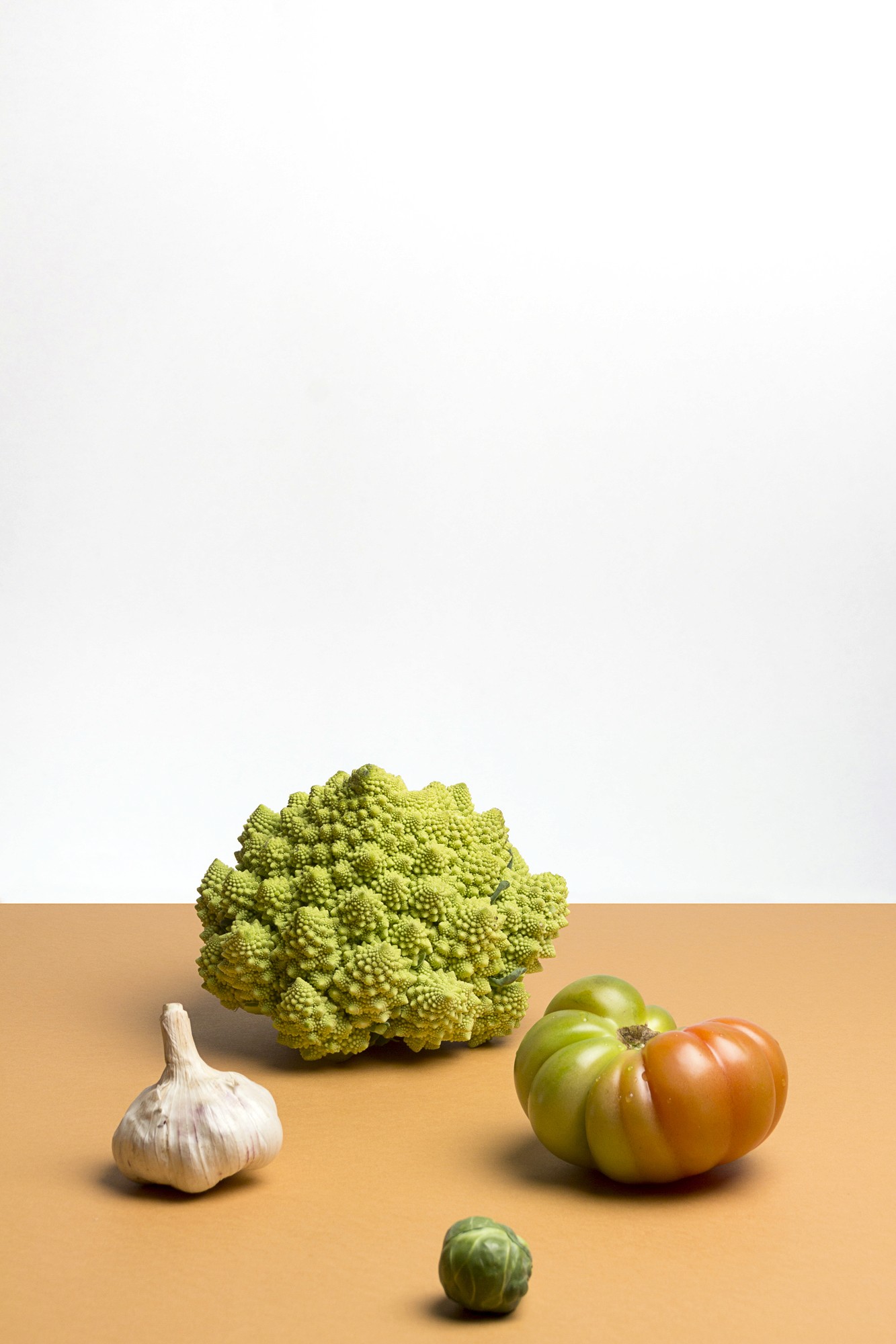 Photo sutudio sur table avec un fond orange d'ingrédients comme un choufleur, une tomate coeur de boeuf, un choux de bruxelles et une gousse d'ail