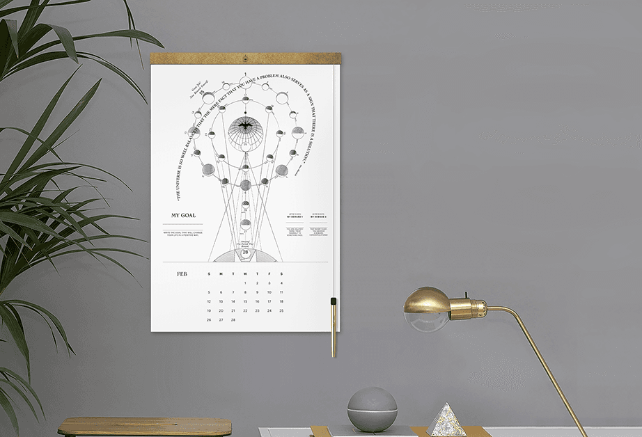 A framed black and white diagram titled “MY GOAL” hangs on a gray wall. The diagram features a central circular design with radiating lines and smaller circles, labeled “MY GOAL” at the top and “FEB” below. Below the diagram is a calendar page showing dates from February to December. To the right, a gold desk lamp with a clear shade sits on a surface next to a gray sphere and a small triangular object. A green plant is partially visible to the left of the frame.
