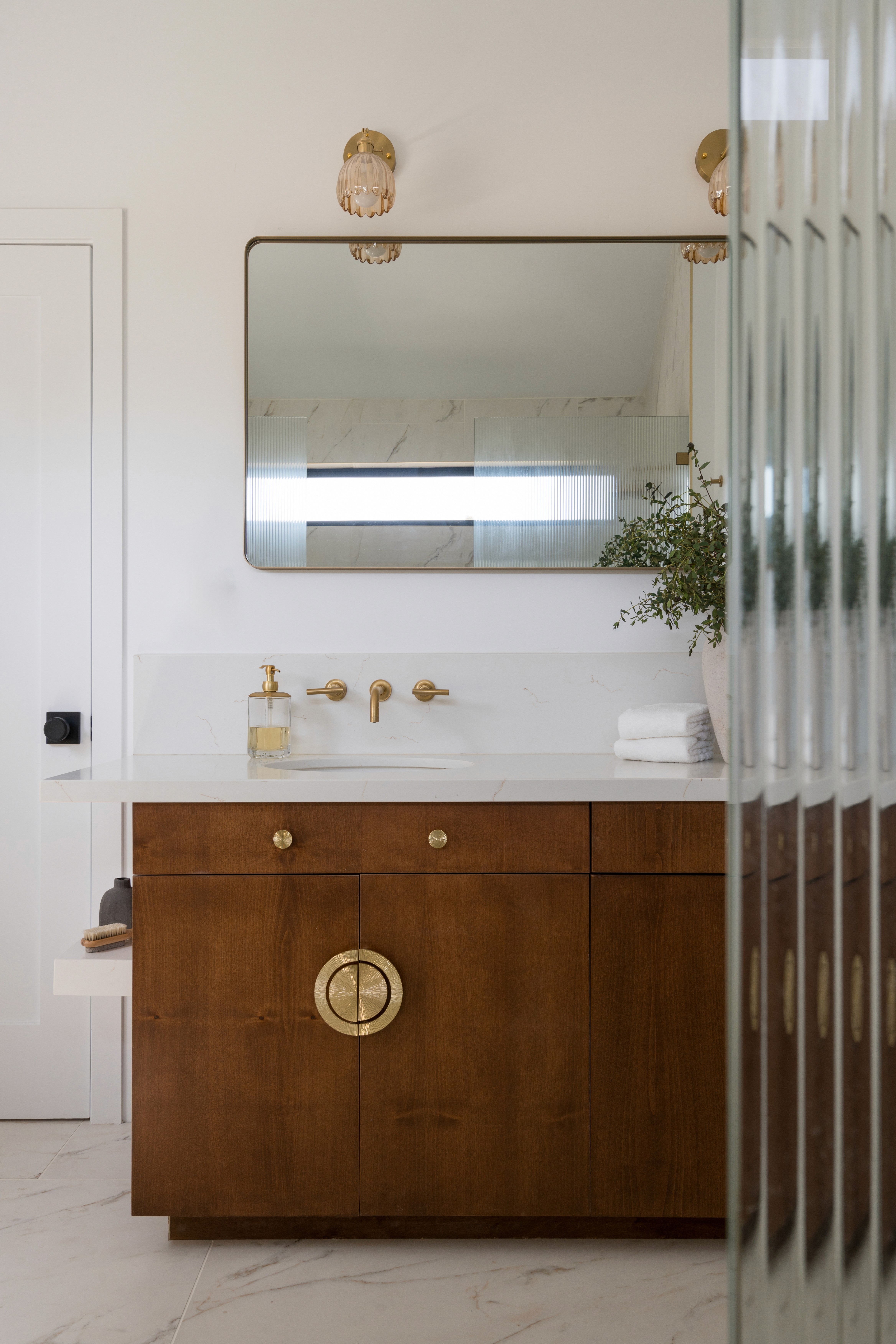 A luxurious primary bathroom adorned with marble accents, a double vanity with modern fixtures, and a glass-enclosed rainfall shower. Soft neutral tones and elegant finishes create a serene, spa-like atmosphere, perfect for relaxation by Jubilee Interiors in Lakewood, California