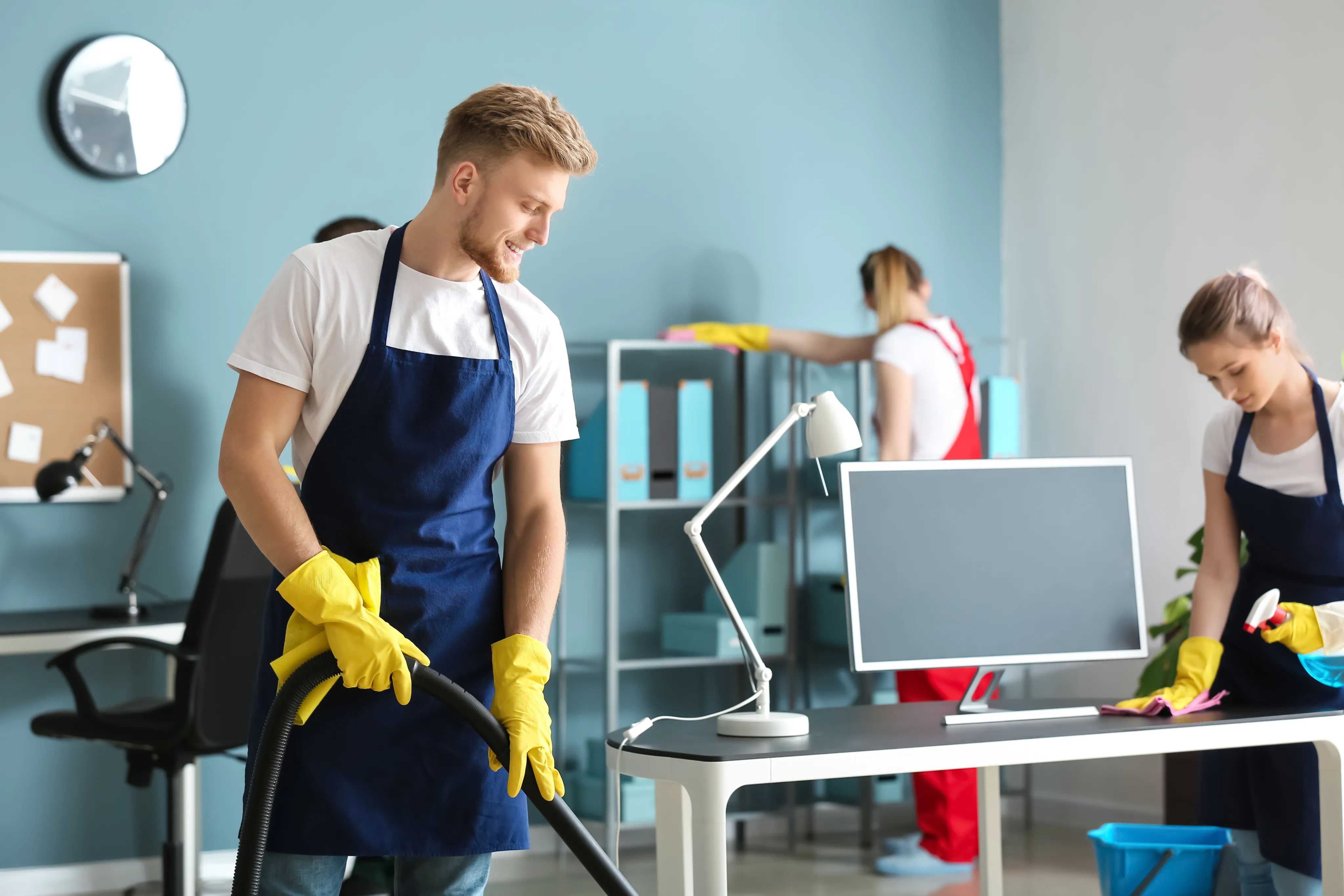 Professional male cleaner in a blue apron and yellow gloves using a vacuum cleaner on a carpet in a bright office setting.