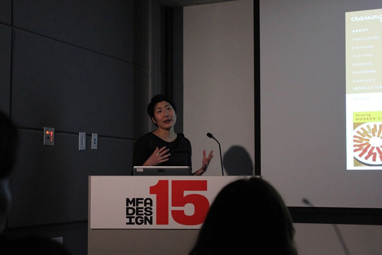 A photo of a woman giving a lecture while sitting at a stand with text MFA DESIGN 15 on it.