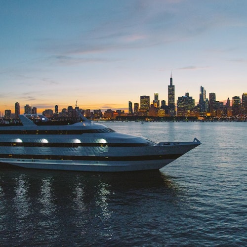 Isang malaking yate ang nakadaong sa kalmadong tubig sa paglubog ng araw, na may tanawin ng lungsod na may matataas na gusali sa likuran.