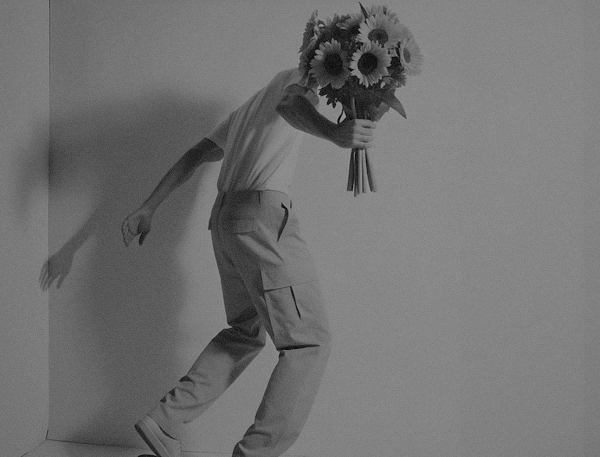 A person skateboards indoors, holding a bouquet of sunflowers, wearing beige cargo pants and sneakers, against a neutral background.