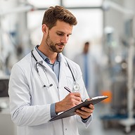 The medical professional sits in the center of a modern practice and documents patient data, with medical equipment in the background.