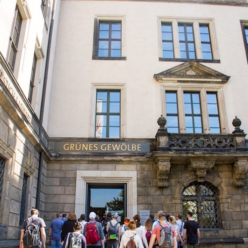 Ihmisiä astumassa sisään rakennukseen, jonka sisäänkäynnin yläpuolella on "GRÜNES GEWÖLBE" -kyltti, suurilla ikkunoilla ja historiallisella arkkitehtuurilla varustettuna.