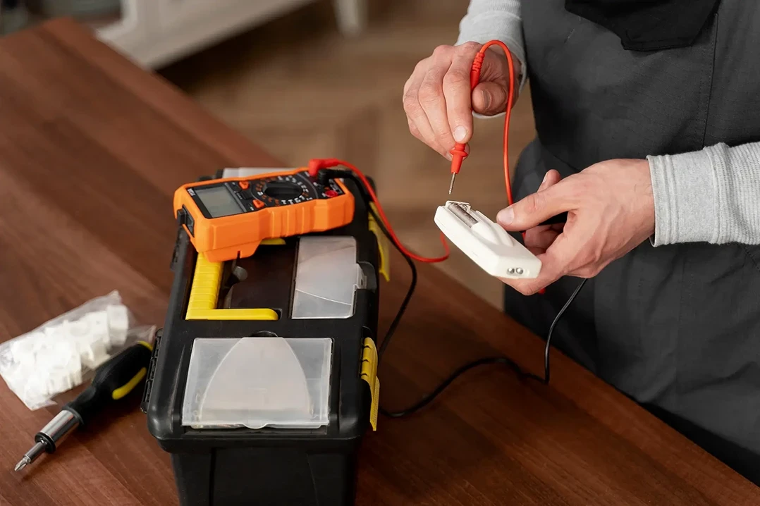 A person uses a multimeter on a wall outlet controller beside a toolbox on a wooden table, conveying focus on home electrical repairs.