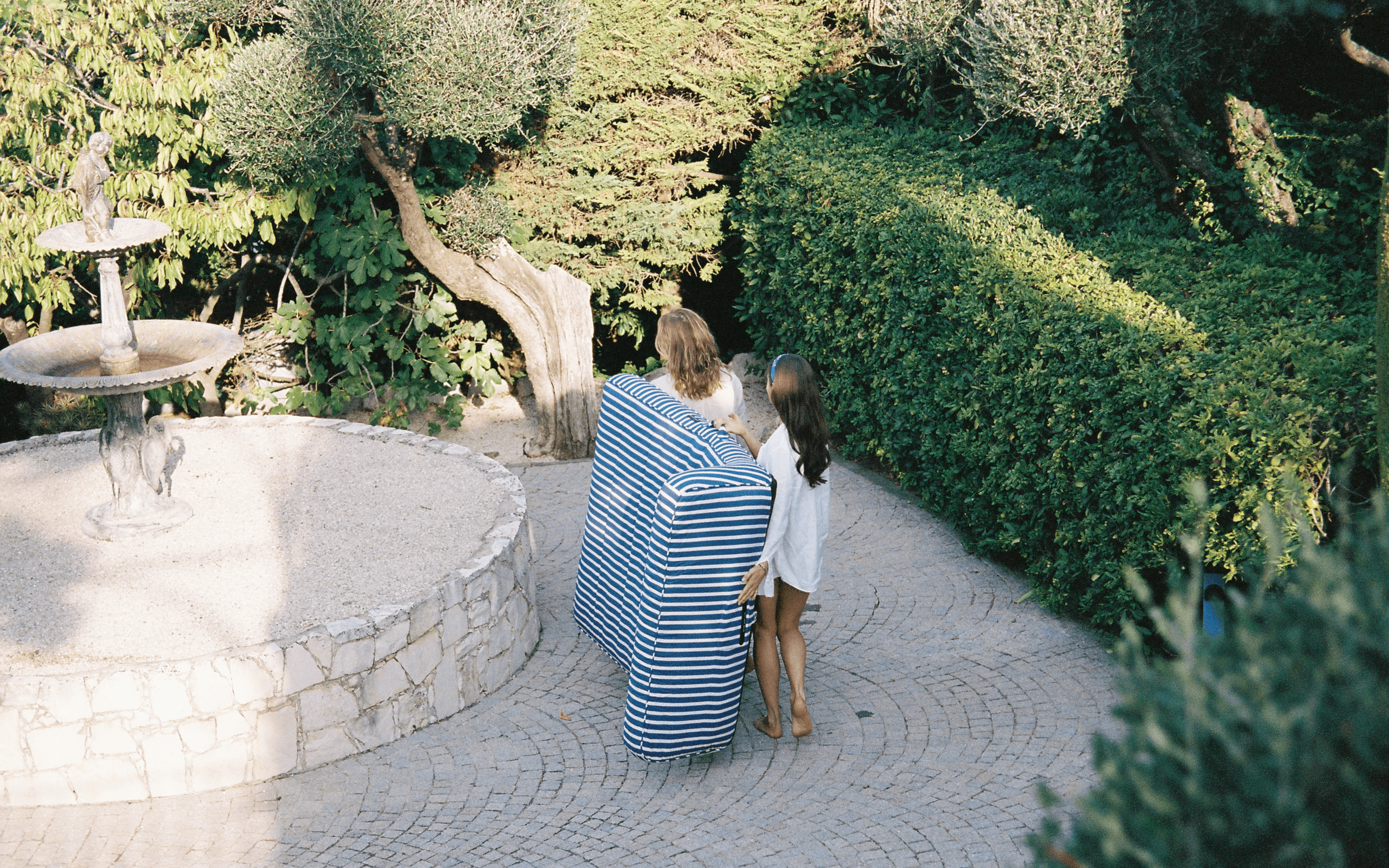 Couple carrying a blue-and-white striped luxury pool float through a lush garden with stone fountain