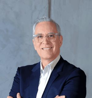A confident-looking person with silver hair and glasses is smiling, dressed in a smart navy blue blazer and white shirt, standing against a modern, light gray textured wall.