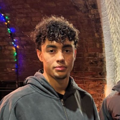 Wolfiez’s X profile avatar: young man in a gray hoodie standing before a brick wall lit by colored lights.