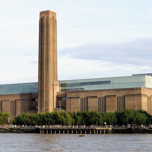 Großes Industriegebäude mit einem hohen zentralen Schornstein, das an einem Fluss liegt, mit Bäumen und einem Spazierweg im Vordergrund.