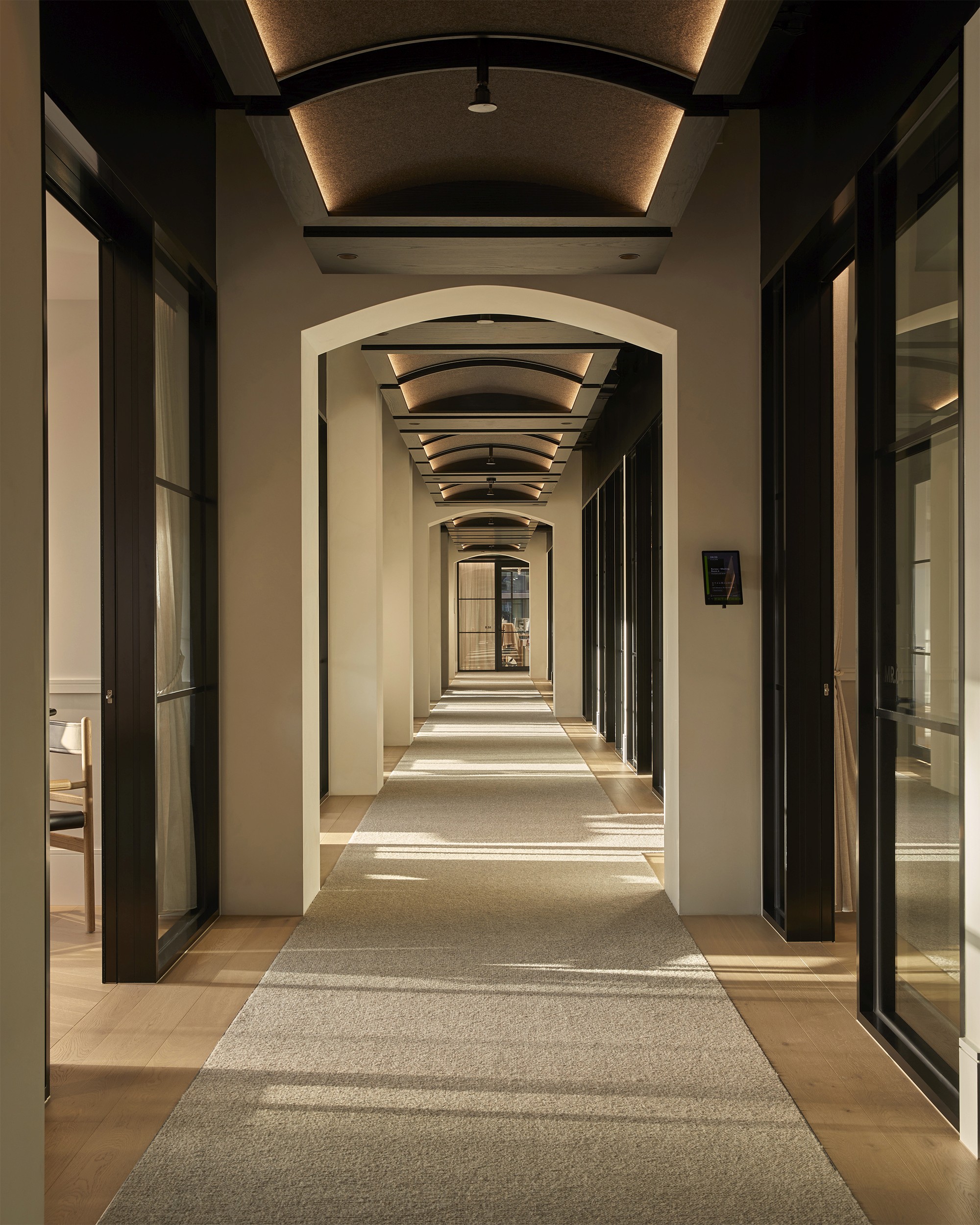 Long arched corridor lined with glass-walled private offices, warm timber flooring and a runner carpet leading toward a window