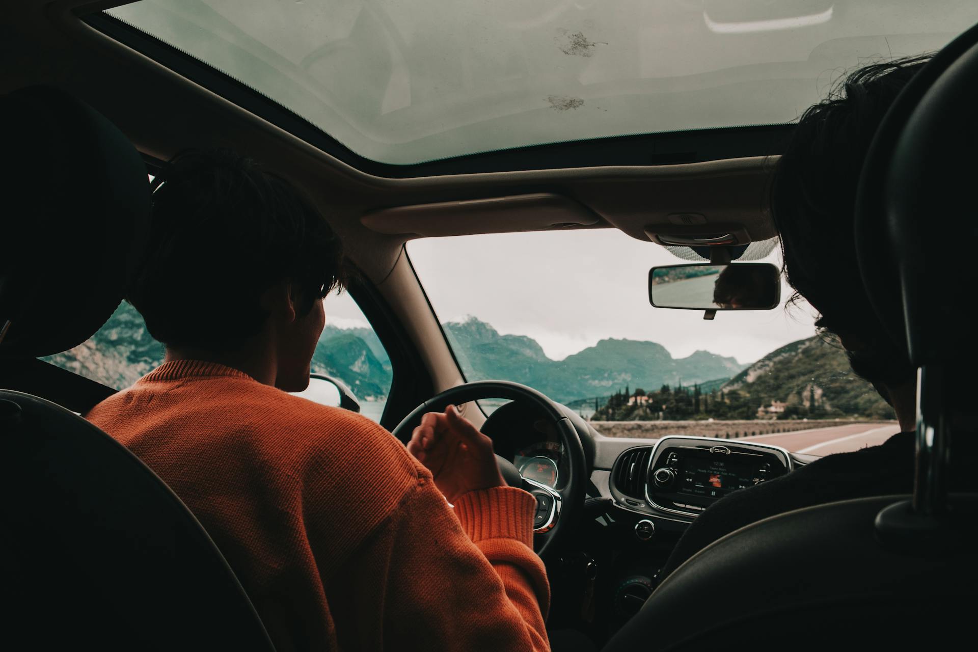 A person with an orange knitted sweater is driving a car with a large sunroof, seen from the back seat. The person's hands are lightly gripping the steering wheel. Through the front windshield, a view of mountains, trees, and a body of water (likely a lake or ocean) is visible under an overcast sky.