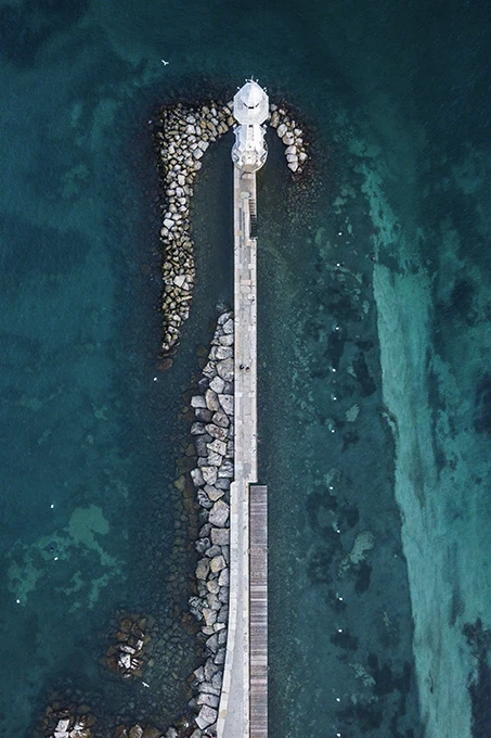 Widok z lotu ptaka na falochron prowadzący do latarni morskiej, otoczony turkusową wodą i kamiennymi umocnieniami