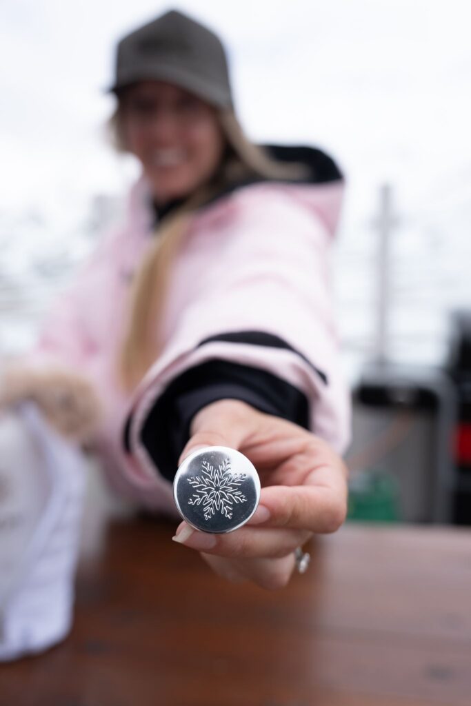 Guest in a pink winter jacket holding a silver bottle cap with an engraved snowflake toward the camera