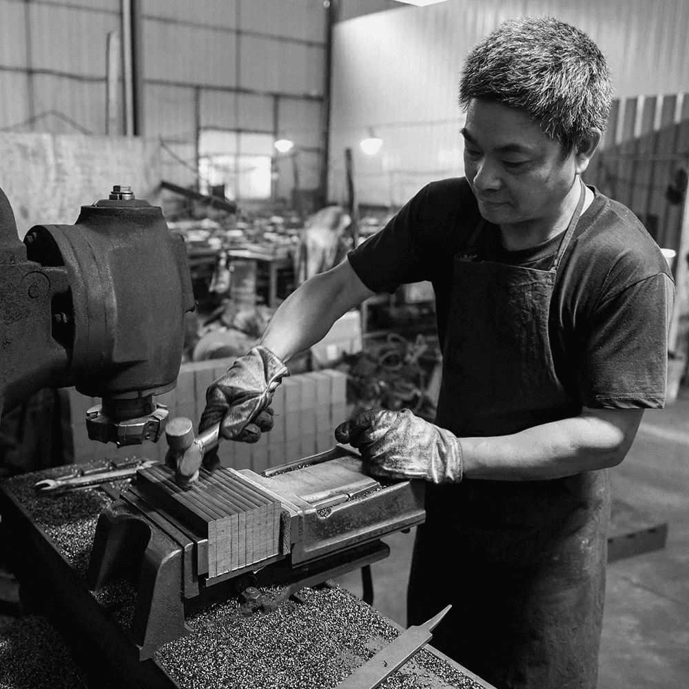 Black and white candid shot of a skilled factory worker in an apron using a hammer and vice to manually calibrate metal components in a furniture manufacturing workshop.