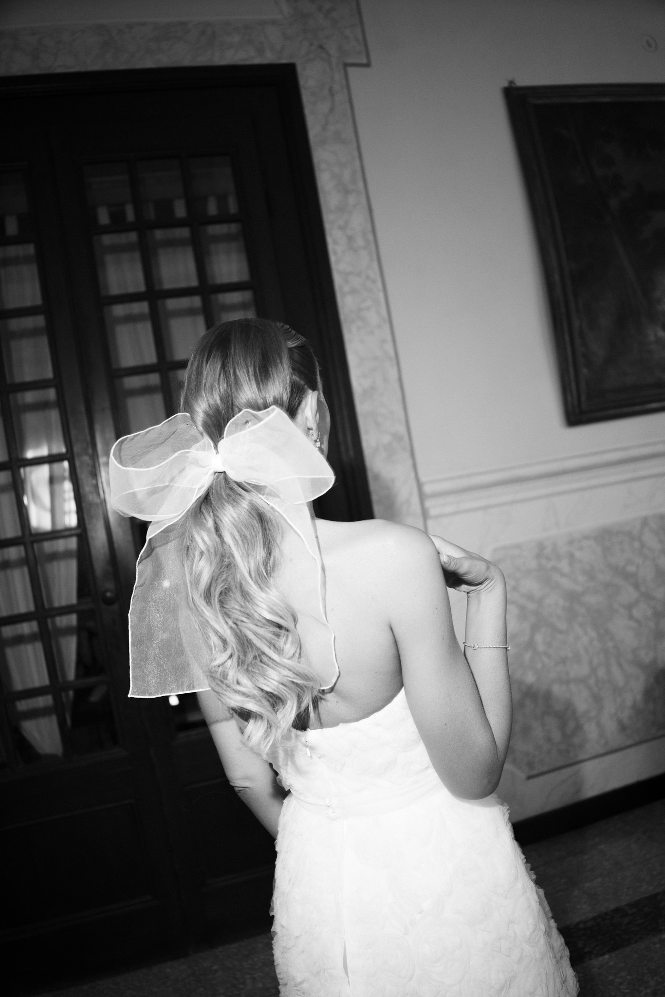 Sposa di spalle con velo bianco annodato, interni Villa Ferrari in bianco e nero, preparativi matrimonio