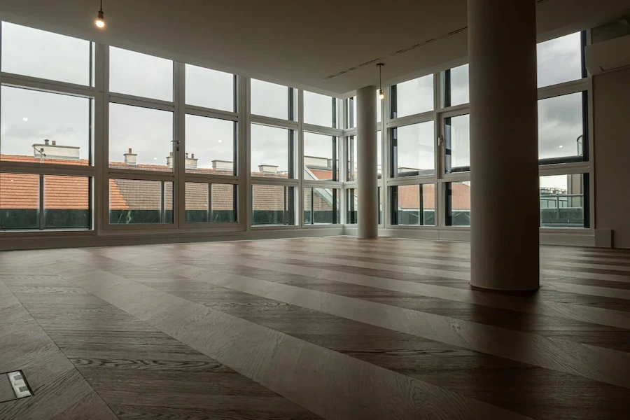 Großer, leerer Innenraum mit bodentiefen Fenstern und frisch verlegtem Parkettboden; moderne, helle Raumgestaltung