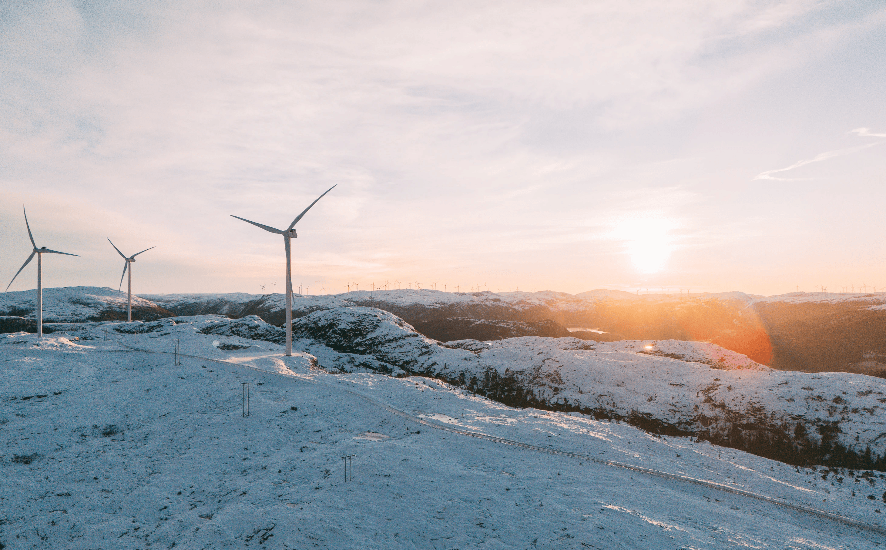 Oversiktsbilde av vindpark med fokus på tre vindturbiner i snødekt mark med solnedgang bak