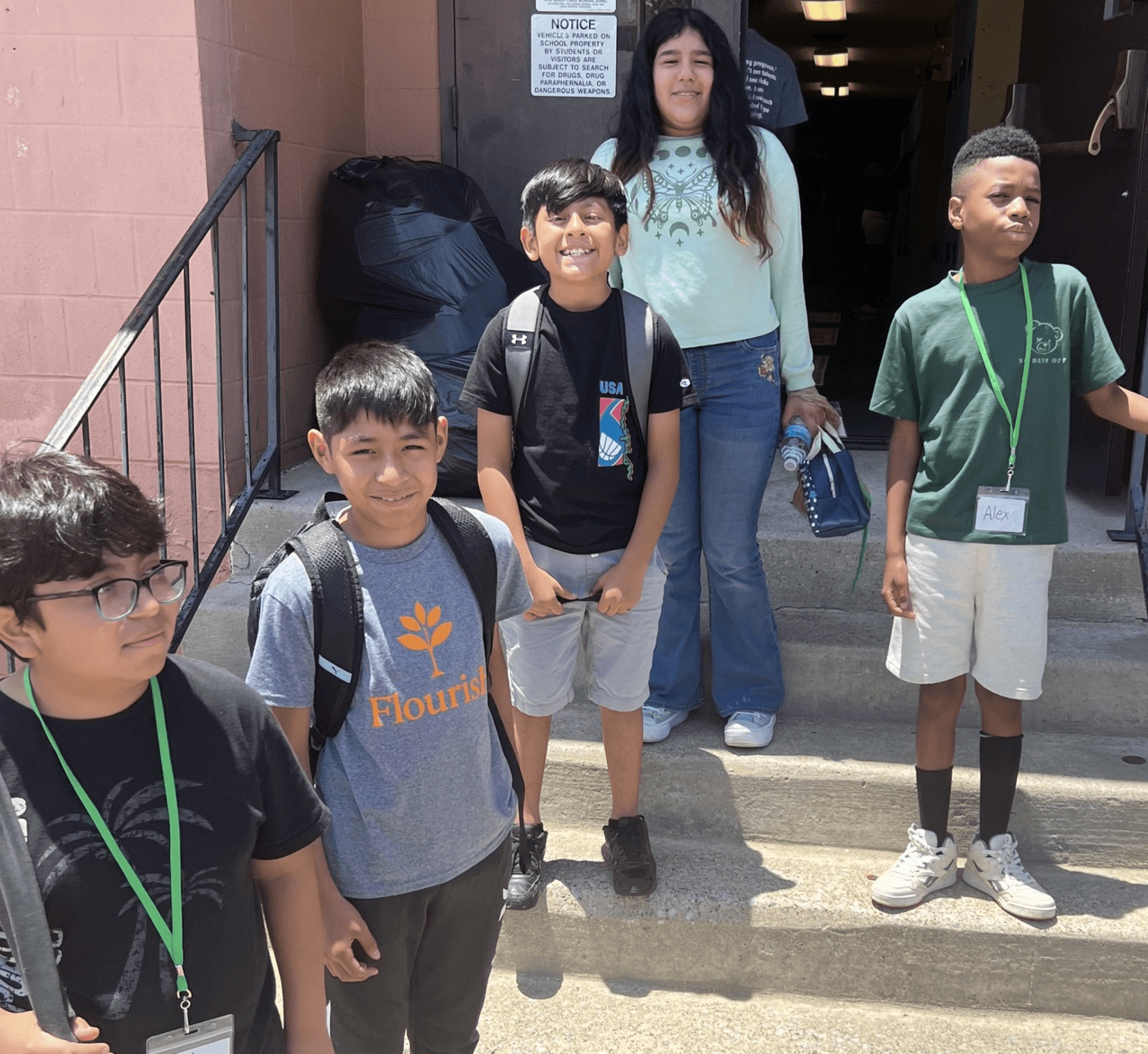 A group of five children stands on steps outside a building, with one adult behind them.