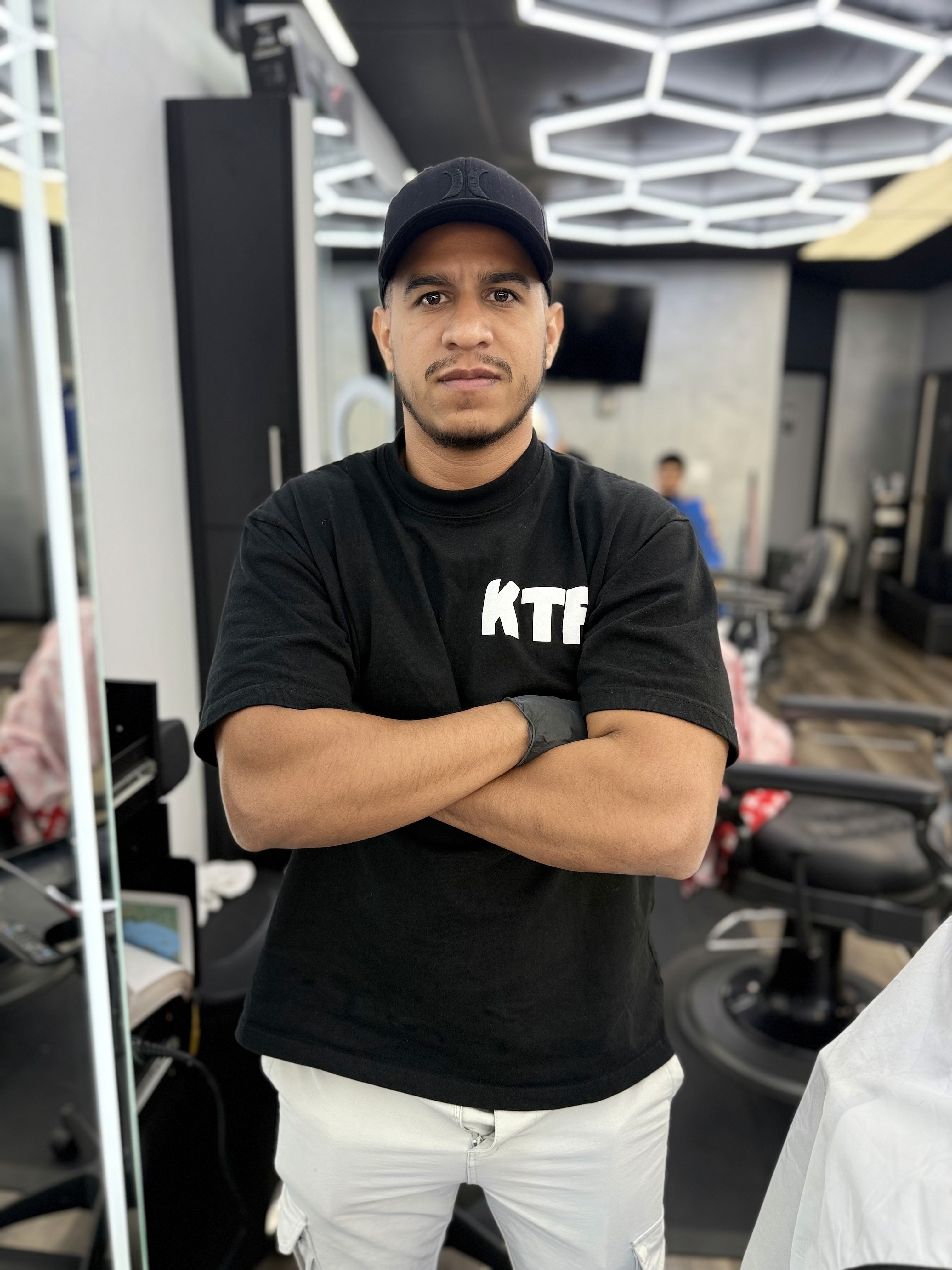 Barber standing with arms crossed in a modern barbershop, wearing a black cap, black T-shirt, and white pants, with hexagon ceiling lights in the background.