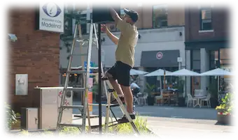 Personne montée sur une échelle installant un haut-parleur dans un espace urbain animé, devant des commerces et terrasses.