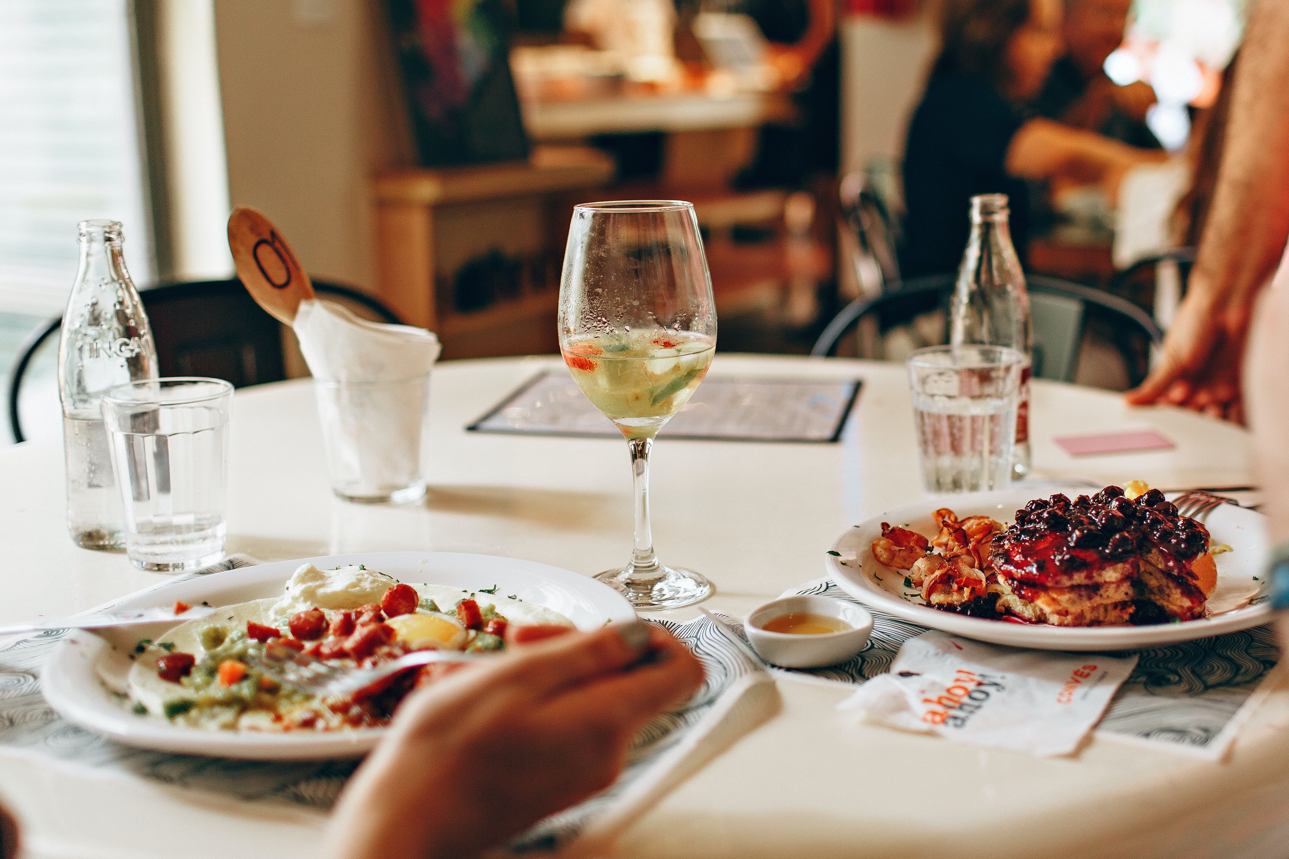 Een tafel gedekt met twee borden eten, een wijnglas en waterglazen, met dinergasten op de achtergrond.