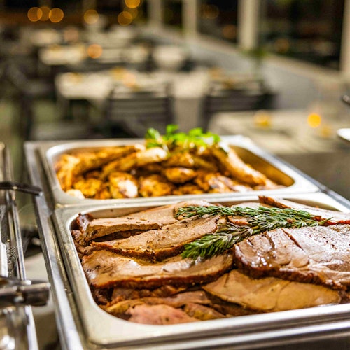 Two trays of sliced meats, one garnished with herbs, in a buffet setting with blurred dining area in the background.