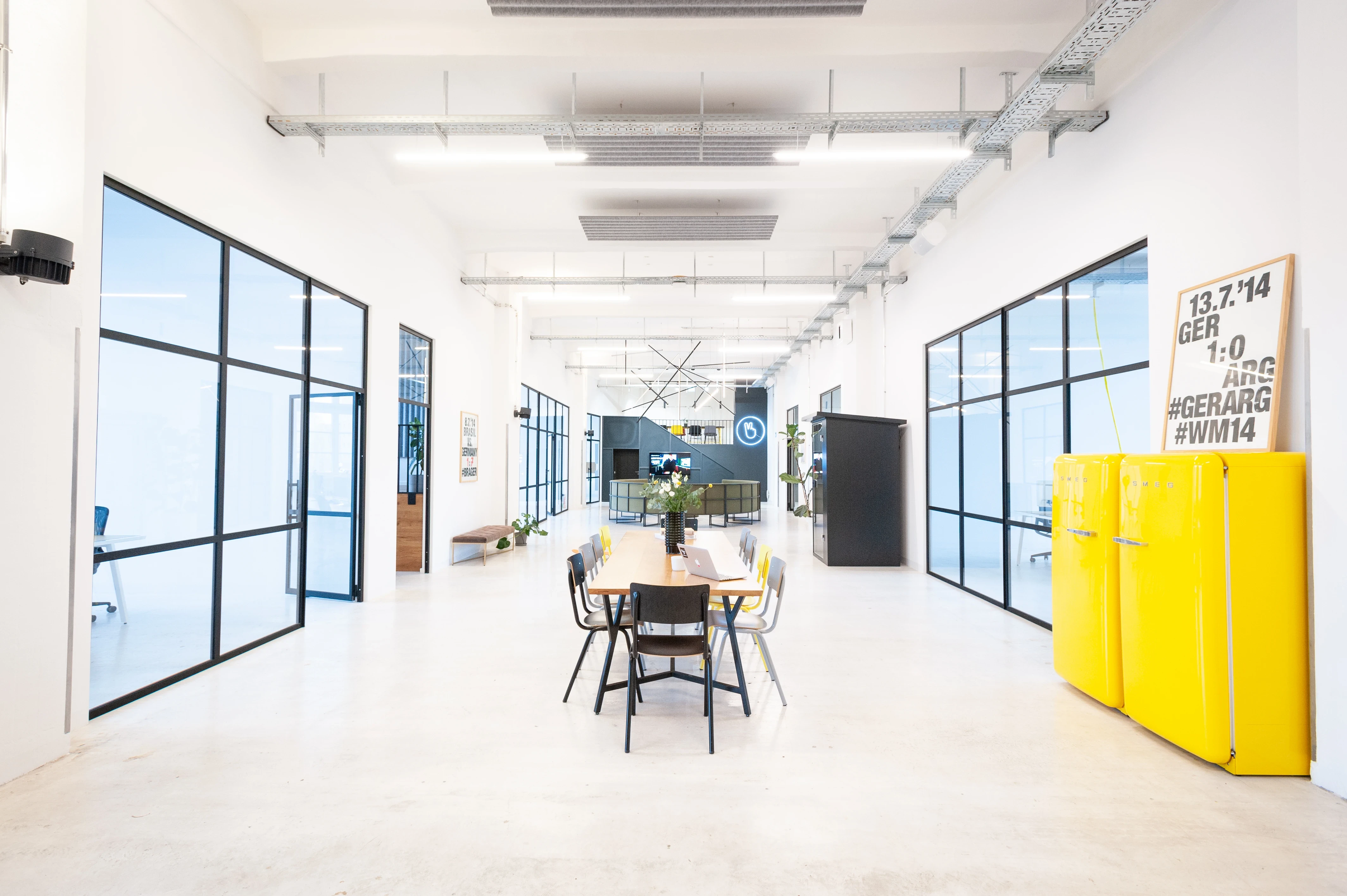 Long open office space at LA RED with white walls, polished concrete floor, black-framed glass partitions on both sides, a central dining table and two bright yellow retro fridges with a World Cup 2014 poster.