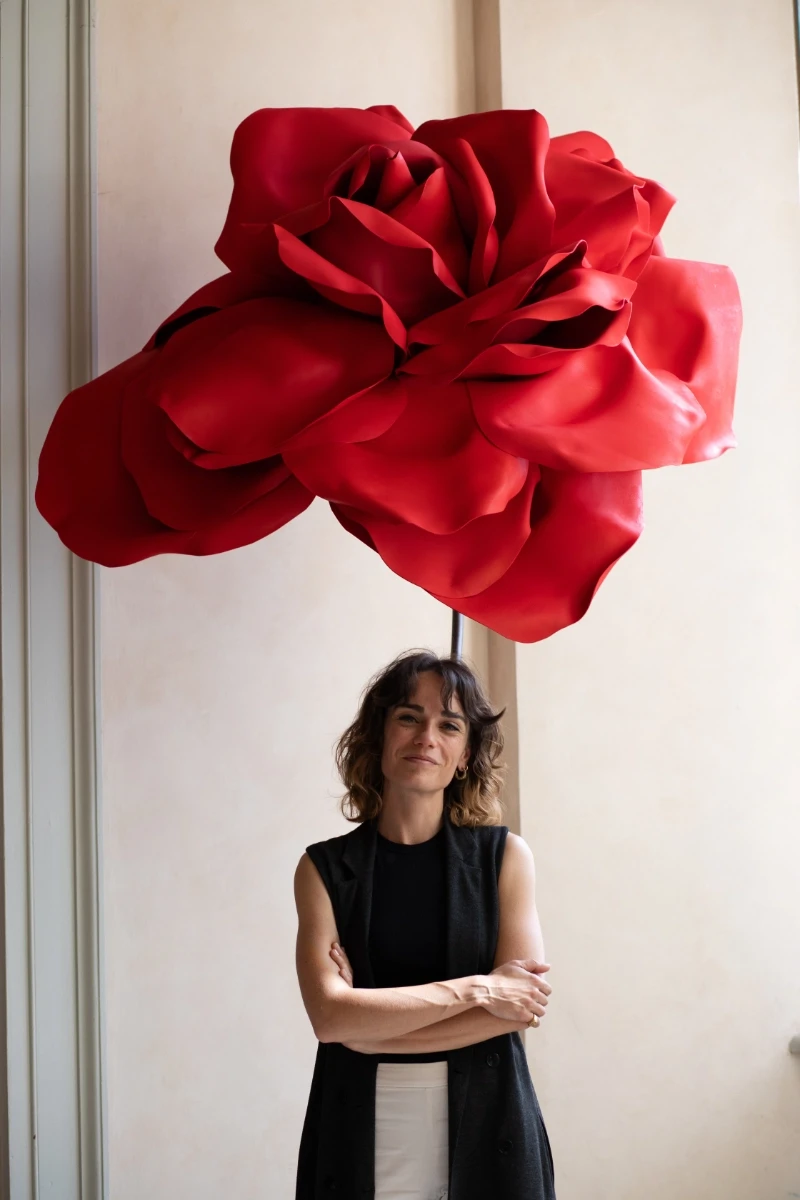 Giulia Caffaro con un abito nero in piedi contro una parete, un grande fiore decorativo rosso dietro di lei.