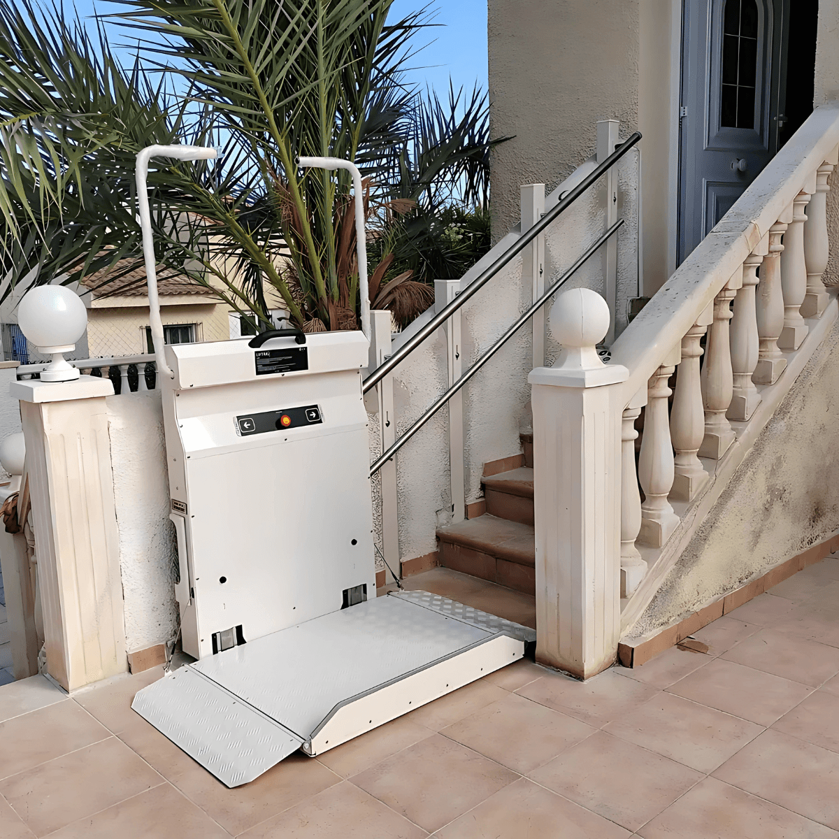 Incline platform stairlift in open deployed position at the base of an outdoor residential staircase — white platform with anti-slip chequer plate surface, control panel with directional buttons, and white balustrade with ornamental pillars