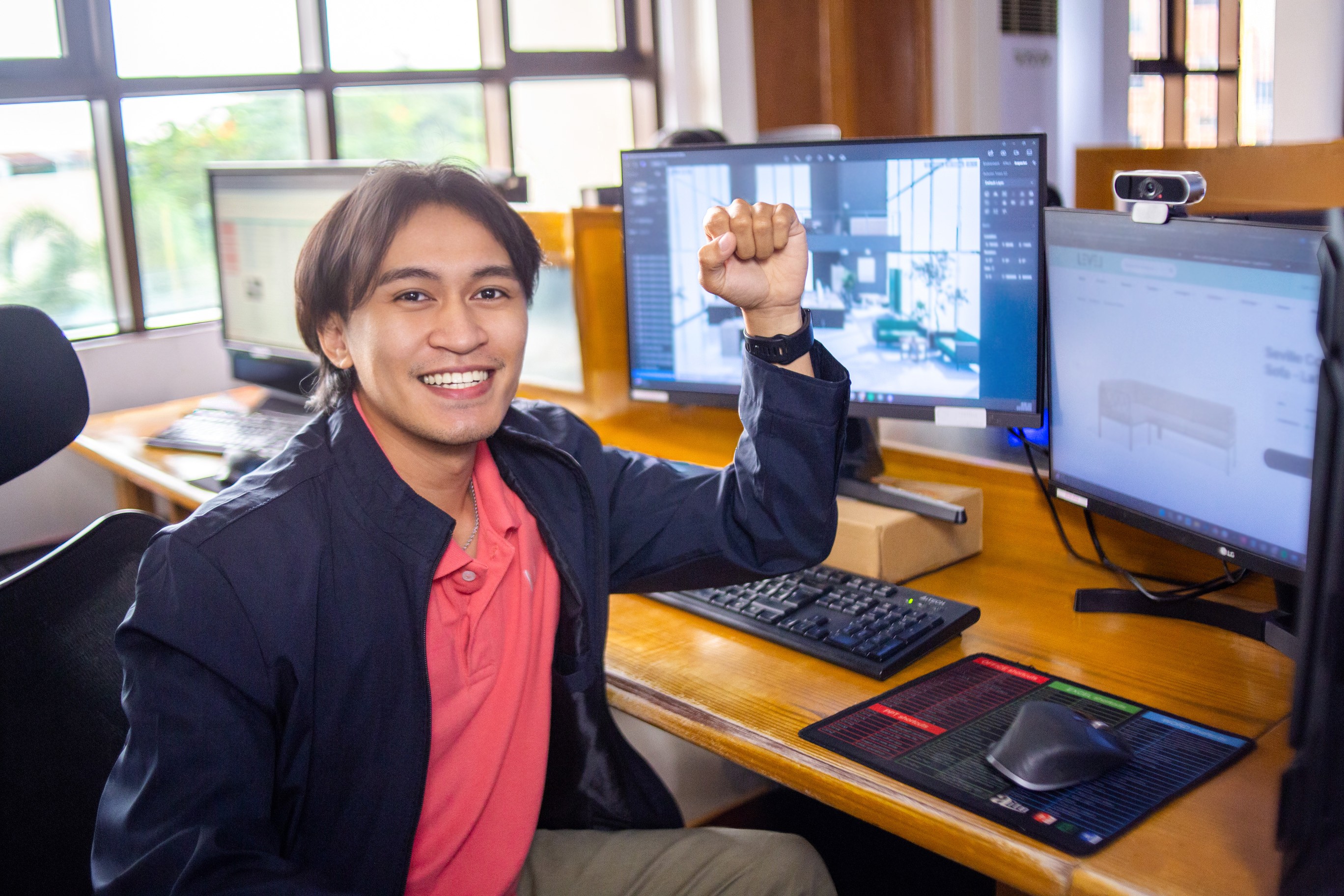 A smiling man gestures energetically while sitting at a dual-monitor workstation in a creative office.