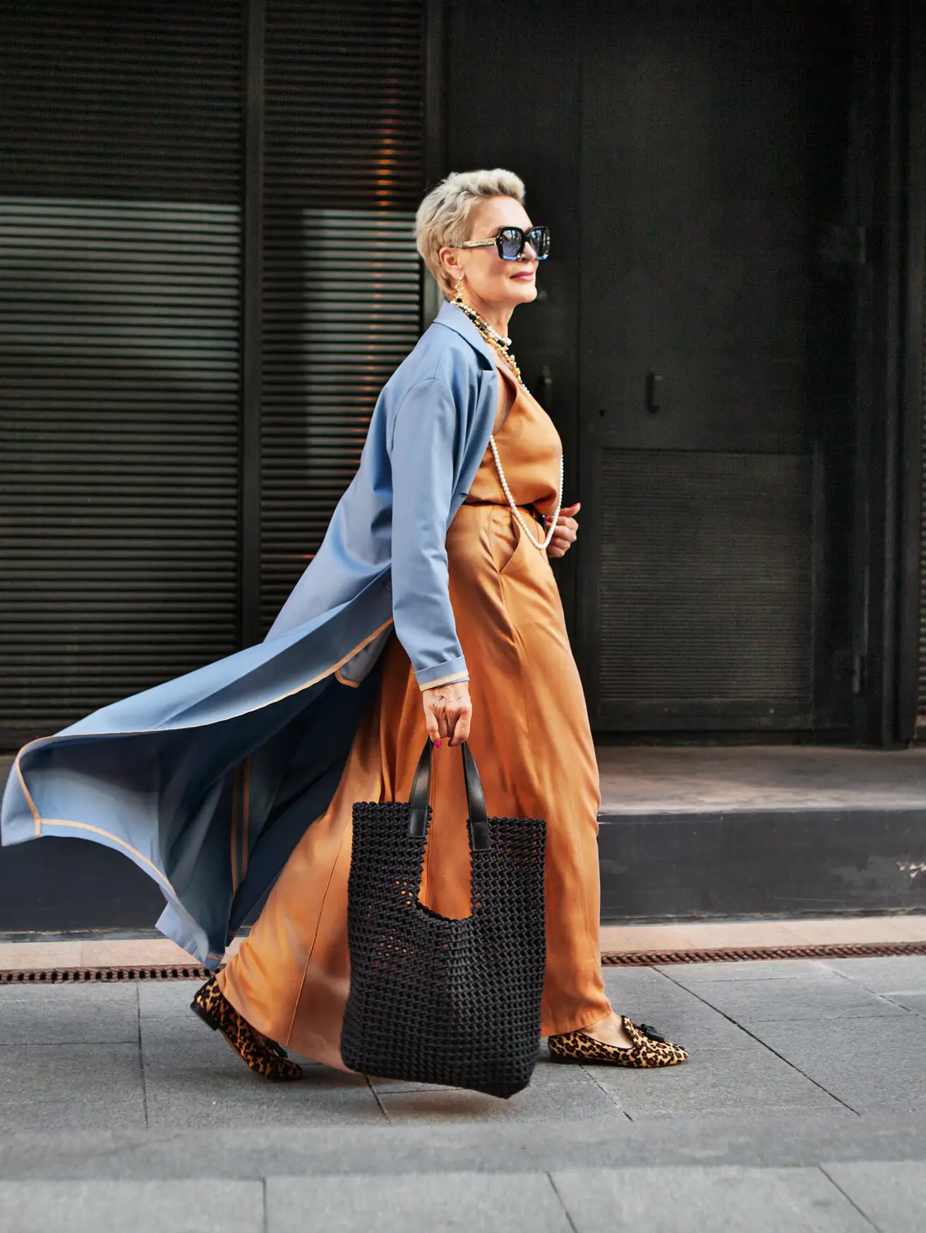 Woman in an amber satin slip dress and pale blue duster coat walking a city street with a black woven tote
