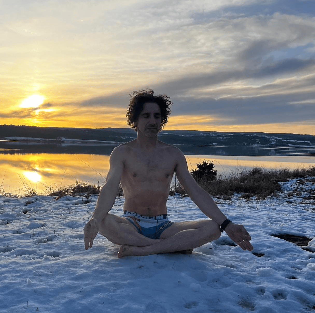 Mathias méditant dans la neige près d’un lac, en sous-vêtements, avec les couleurs du lever du soleil se reflétant sur l’eau.