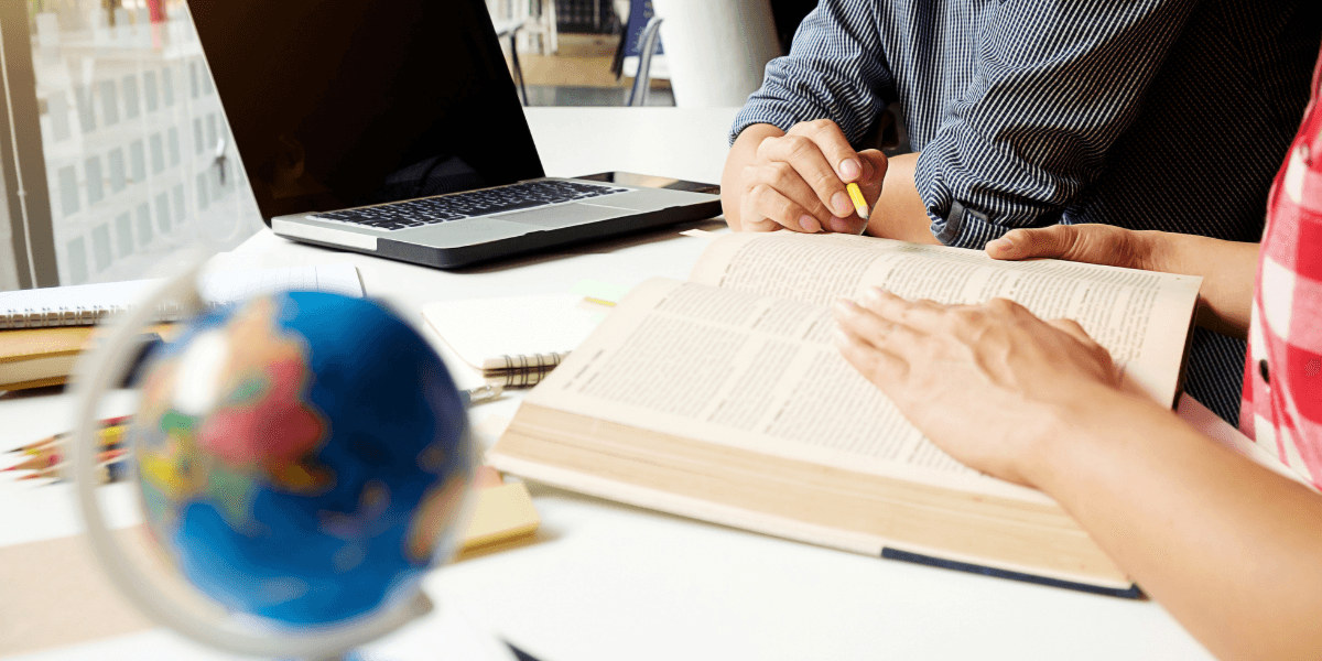 Two people study at a bright desk with an open book and a globe. A laptop is nearby, suggesting a focus on research or learning.