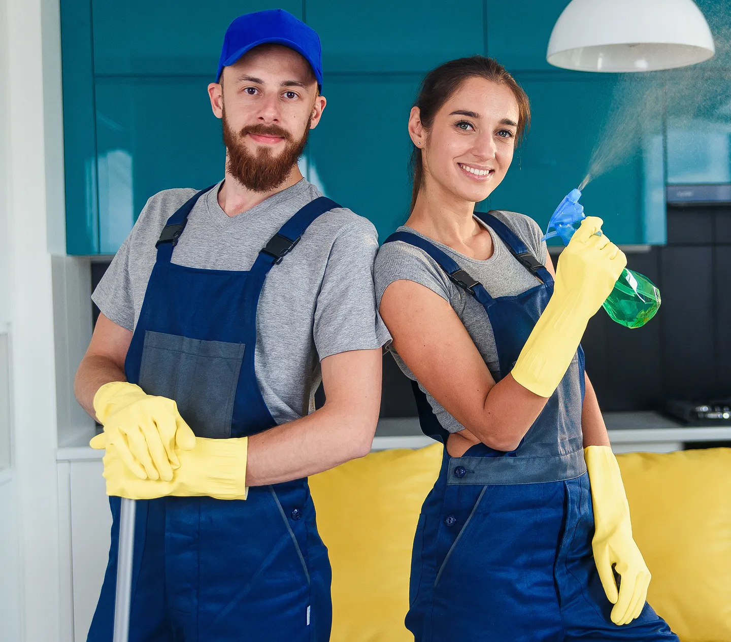 Two professional cleaners, a man and a woman, wearing blue overalls and yellow gloves, posing in a modern kitchen while holding cleaning equipment.