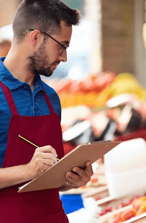 A man in a red apron takes notes at an outdoor fruit stand.