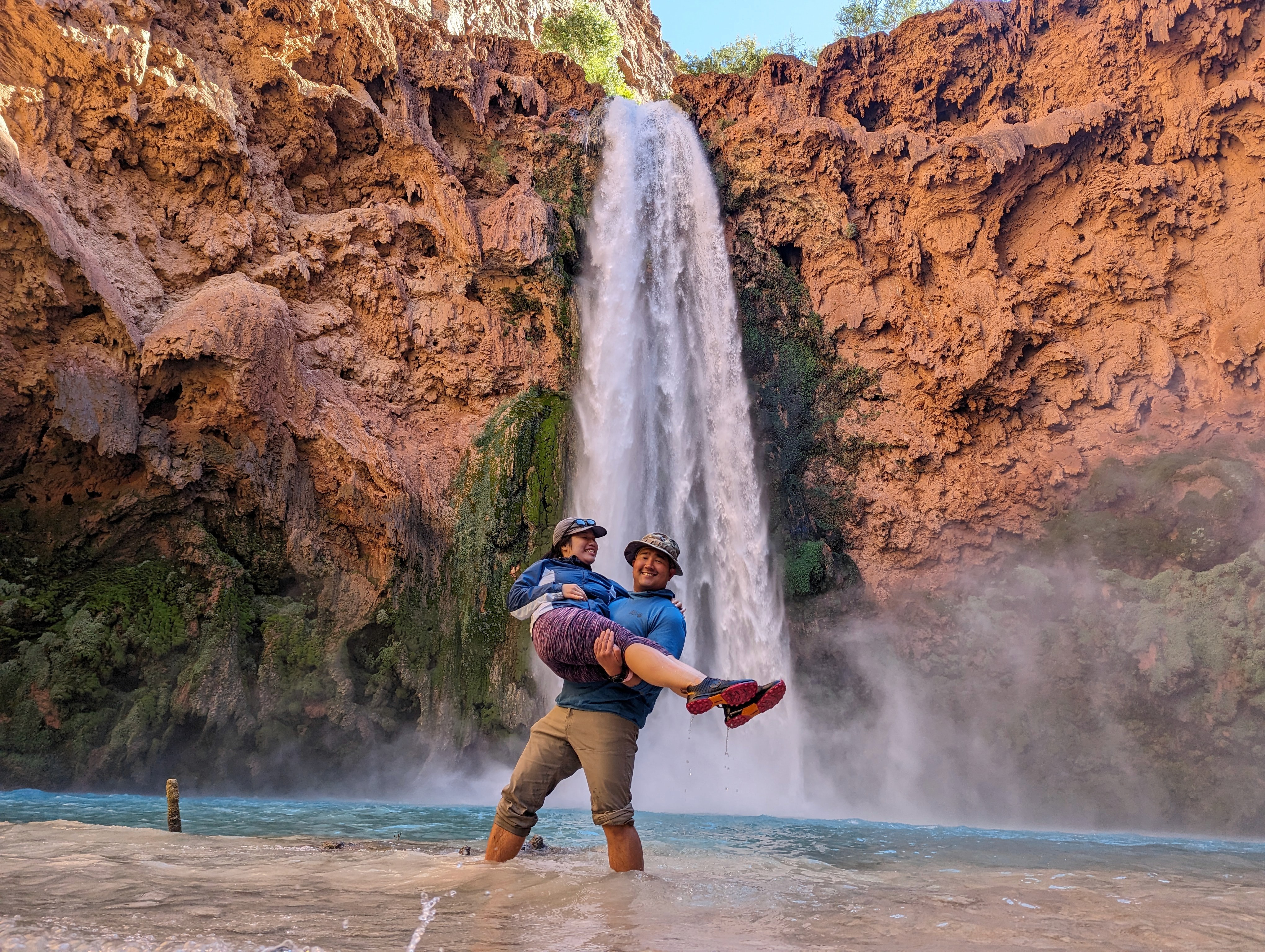 A bride and groom hold hands tenderly, the bride's bouquet of dried flowers hanging low. They stand in a softly lit, elegant interior, conveying romance.