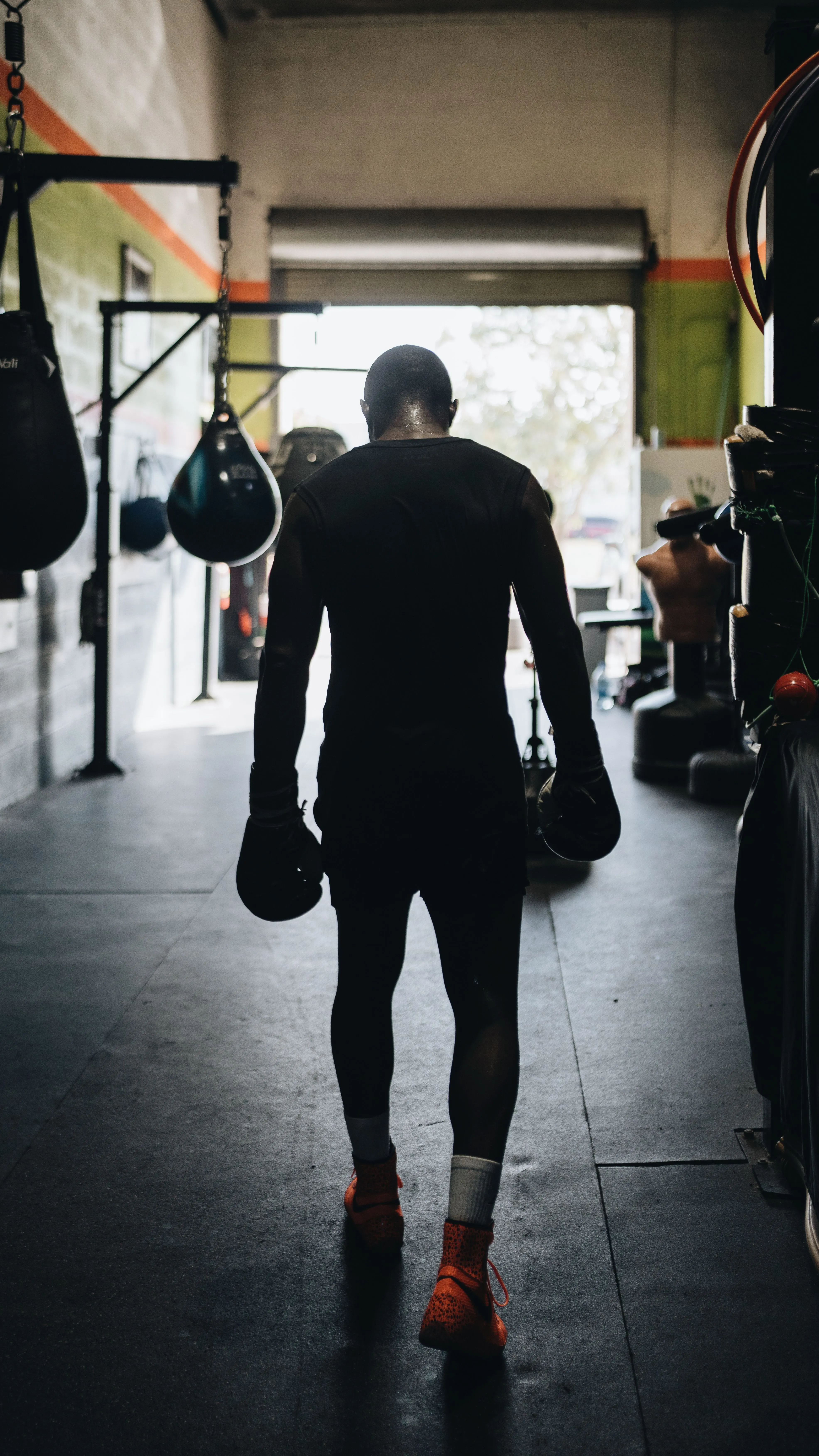 A silhouette of a person wearing boxing gloves and athletic shoes is walking through a dimly lit gym, with punching bags hanging and various exercise equipment visible in the background.
