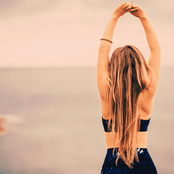 Woman on beach facing the ocean in navy blue yoga clothes with her arms raised over her head stretching